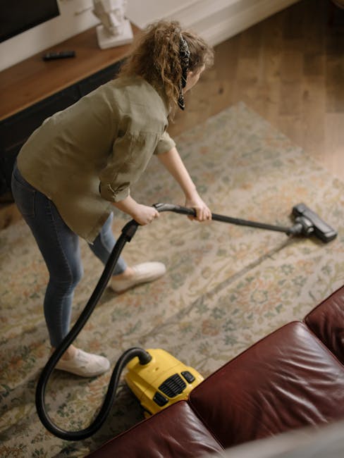 A person holding a yellow portable carpet cleaning machine with a black handle and a black collection tank, positioned on a patterned carpet in a well-lit room. The carpet features a floral or abstrac