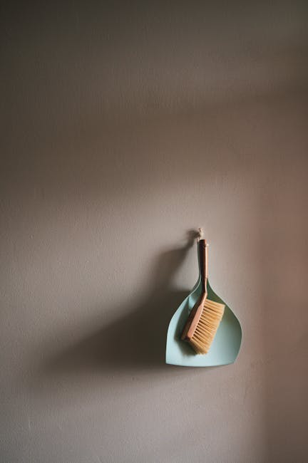 A close-up of a light blue wall hook mounted on a beige wall, holding a wooden-handled scrubbing brush with natural bristles and a light-colored cleaning cloth. The setup suggests a tidy and organized space, likely in a kitchen or utility room, with an emphasis on surface cleaning and maintenance. The lighting is natural, casting subtle shadows, and emphasizes the simplicity and cleanliness of the scene. This image is associated with domestic cleaning and hygiene practices, as promoted by Carpet Cleaning Kentish Town, highlighting preparation for surface sanitisation and deep cleaning routines.