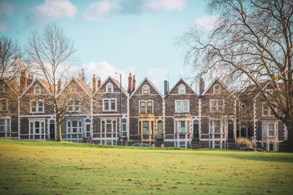 A row of Victorian-style terraced houses with brick and stone facades, large front windows, and pitched roofs, set behind a well-maintained grassy lawn with a few leafless trees. The houses are illuminated by natural daylight, highlighting the cleanliness and neat appearance of the exteriors. This residential setting exemplifies the importance of surface cleaning and maintenance, with visible details such as glass panes in the windows, brickwork, and wooden elements that benefit from regular deep cleaning and sanitisation, which is a service offered by Carpet Cleaning Kentish Town. The scene emphasizes the importance of proper property upkeep and hygiene within domestic environments.