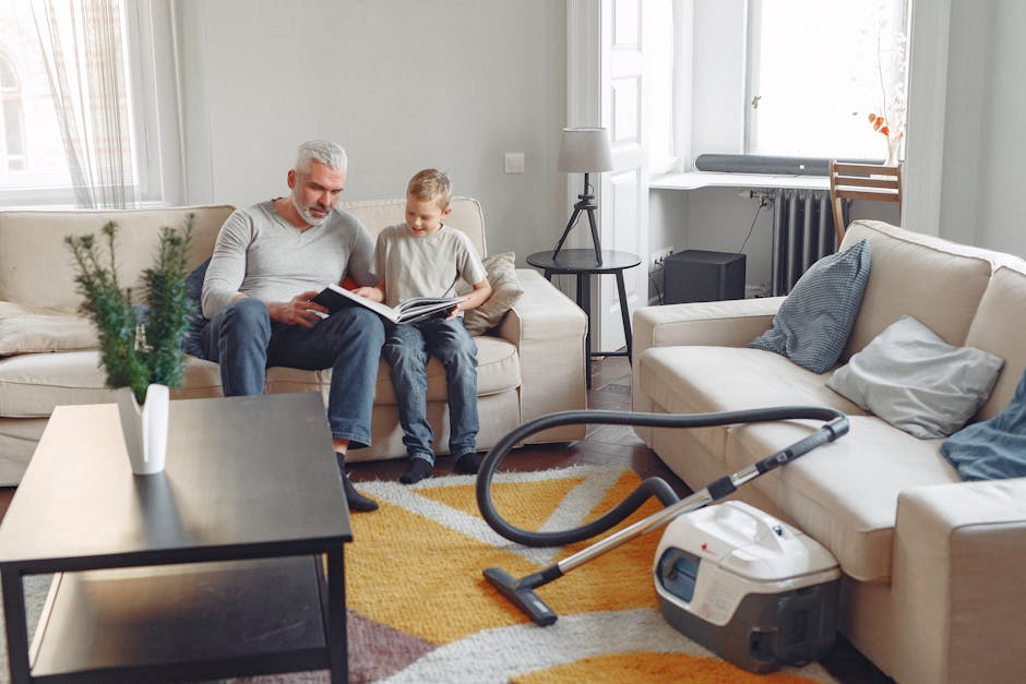 A person kneeling on a patterned carpet in a living room or similar space, using a yellow and black vacuum cleaner for surface cleaning. The individual is holding a white cloth or filter inside the vacuum's open top compartment. The room has a soft natural light, and the carpet displays an intricate floral and geometric pattern. This scene reflects domestic cleaning activities, emphasizing the use of cleaning appliances to maintain hygiene and cleanliness. The background shows a blurred section of the room, suggesting a tidy environment. Carpet Cleaning Kentish Town offers professional deep cleaning services, ensuring effective sanitisation and maintenance of residential surfaces. Visible tools include the vacuum cleaner with a flexible hose attachment, highlighting modern cleaning equipment used for routine cleaning or intensive surface sanitisation.