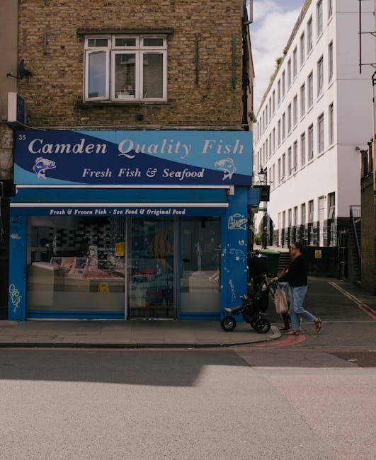 Exterior view of a fish and seafood shop named 'Camden Quality Fish' situated on a brick building, with a blue signage board displaying the shop's name and offerings. The shop features large glass win